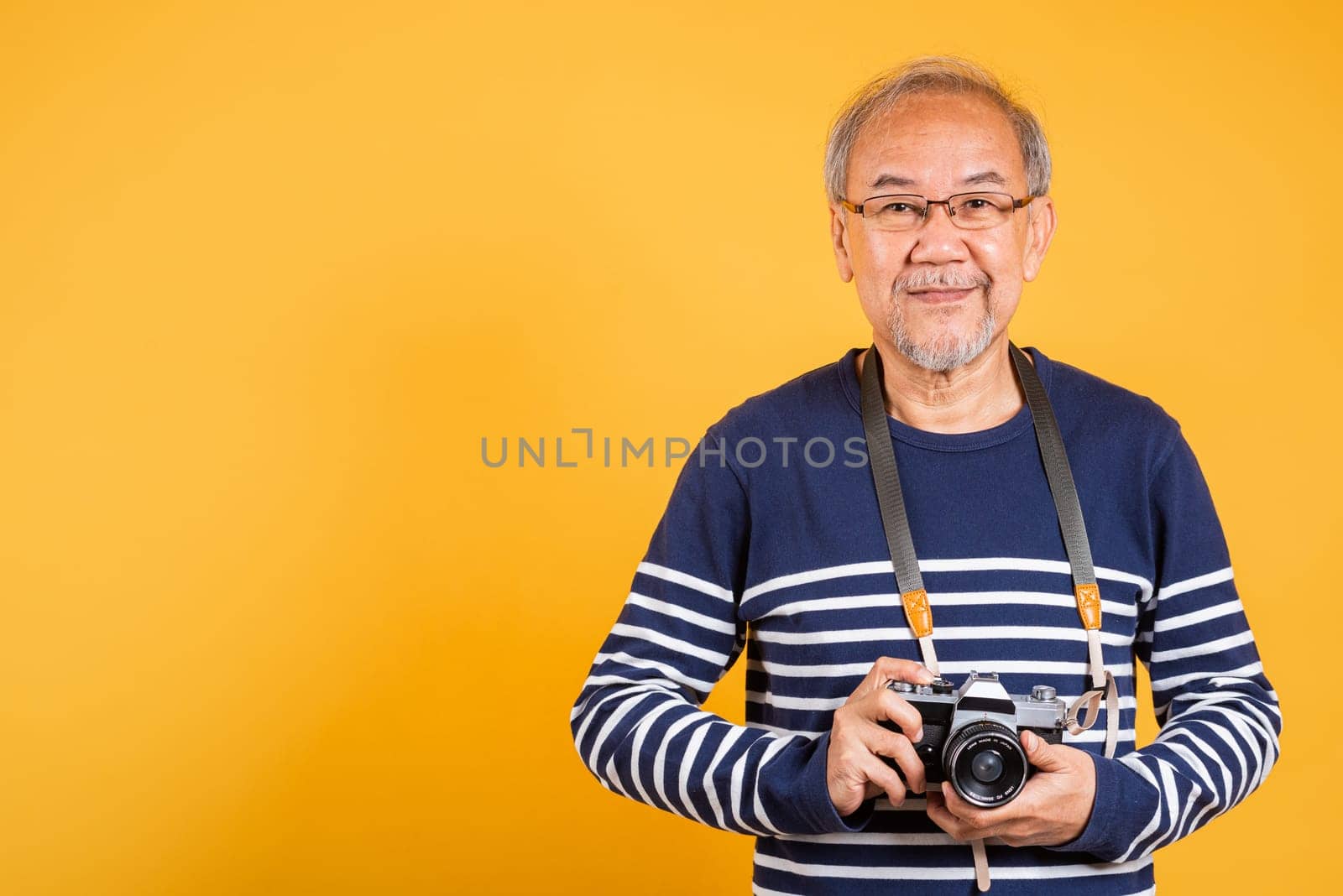 Portrait Asian old man wearing glasses holding vintage SLR camera studio shot isolated yellow background, smiling happy Photographer elderly man gray haired holds old fashioned camera