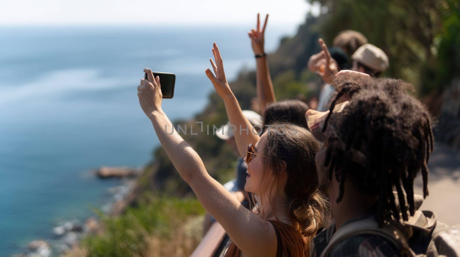 Young diverse friends taking photos and showing peace signs while standing on a picturesque cliff by the sea. Great for vacation themes, youth culture, and outdoor lifestyle content