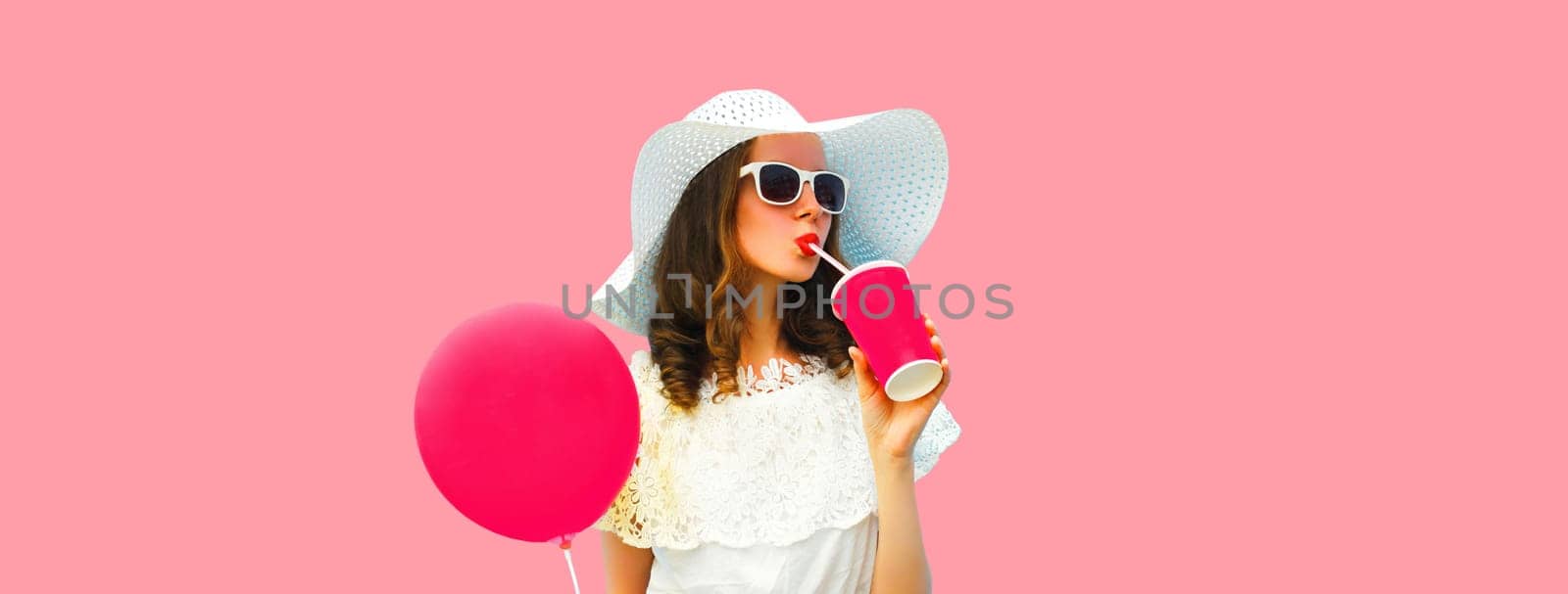 Portrait of beautiful young woman model with cup of coffee or fresh juice holding balloon wearing white summer straw hat on pink studio background
