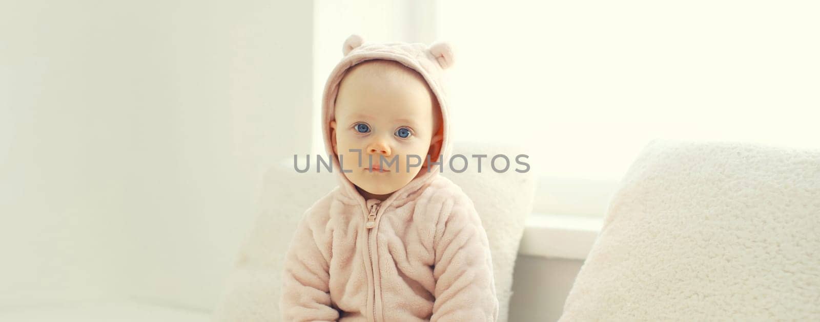 Cute little baby sitting on the floor in bathrobe in white room at home near window