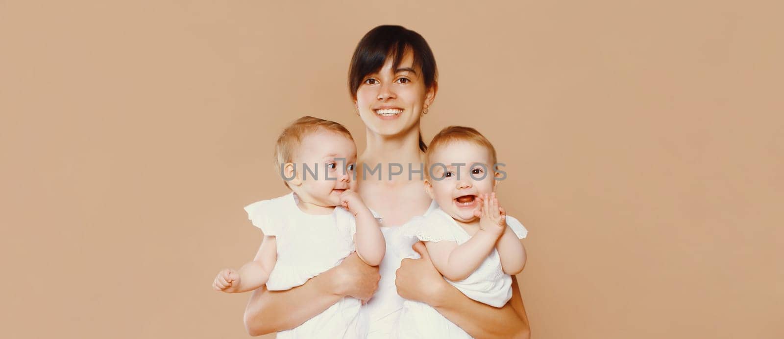 Portrait of happy smiling young mother holding twin babies on brown studio background