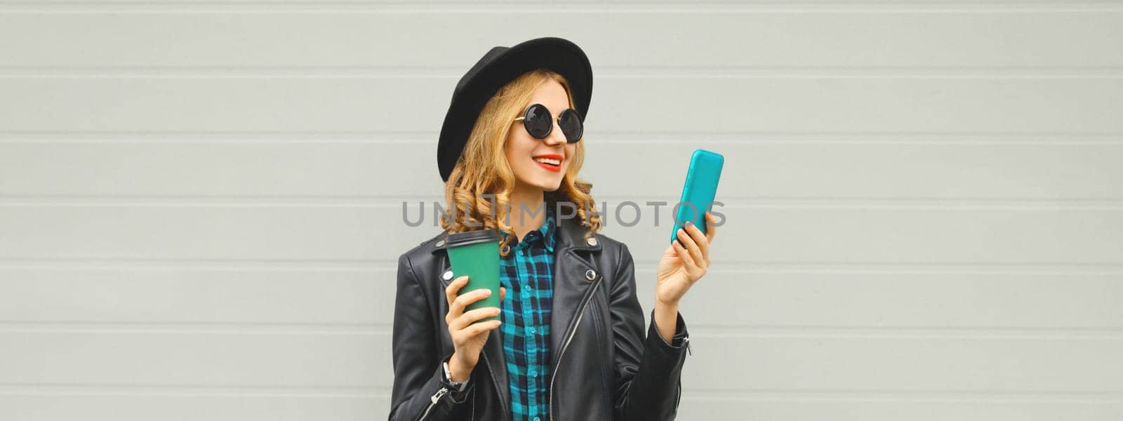 Stylish happy smiling modern young woman with phone, girl holding smartphone, cup of coffee drink in black hat, jacket on gray background