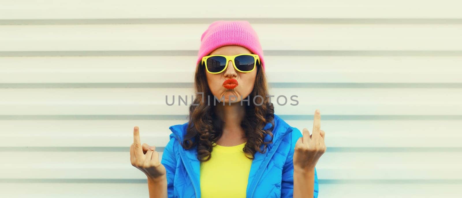 Portrait of bad angry young woman expression showing hand with middle finger sign, stylish girl wearing vivid colorful clothing, pink hat, glasses on white background