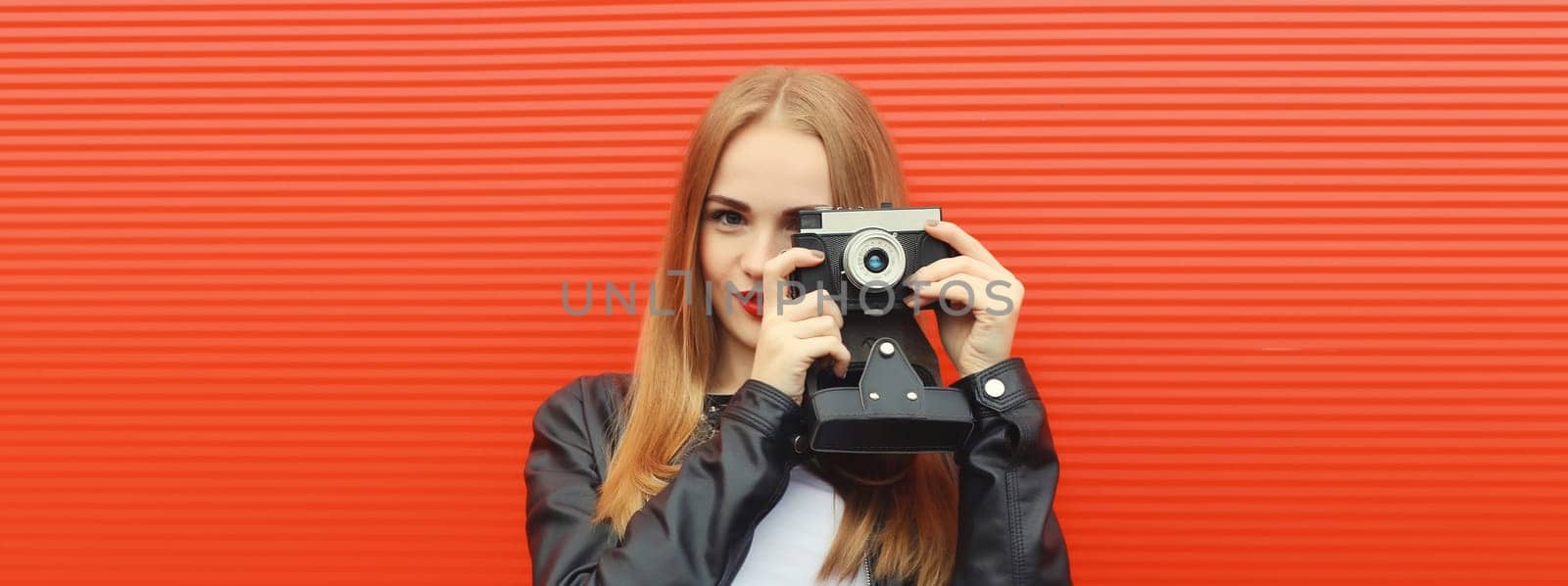Portrait of stylish young woman photographer with film camera, modern girl taking a picture standing on red background