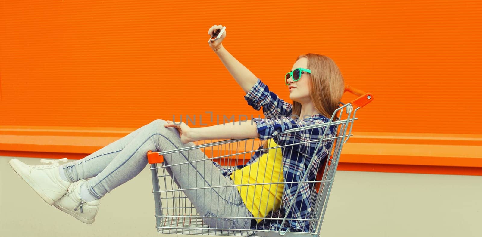 Stylish happy young woman taking selfie with phone, cool modern teenage girl having fun sitting in shopping trolley cart in the city on a summer day