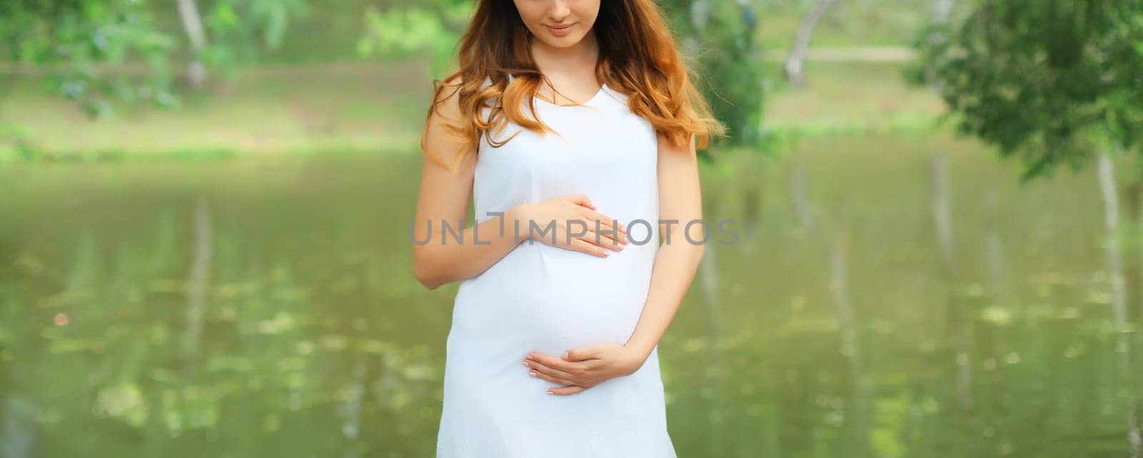 Close-up of happy young pregnant woman walking in summer park against lake background