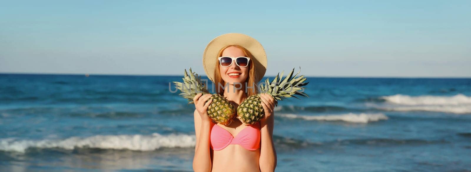 Summer vacation, beautiful happy smiling woman in pink bikini swimsuit, tourist straw hat holding pineapple fruits on the beach on sea coast background