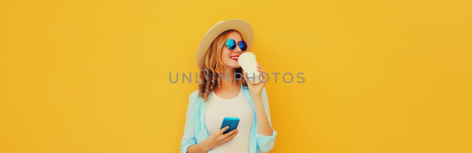 Summer stylish happy young woman drinking coffee with phone, cool girl using smartphone holding cup of coffee dressed tourist straw hat looking away standing on yellow background