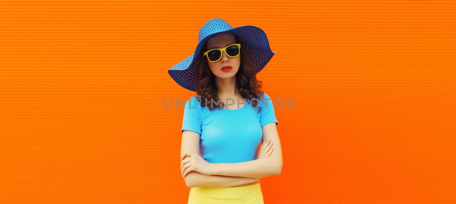 Beautiful stylish young woman, fashionable female model in summer blue straw hat posing on vivid orange wall background