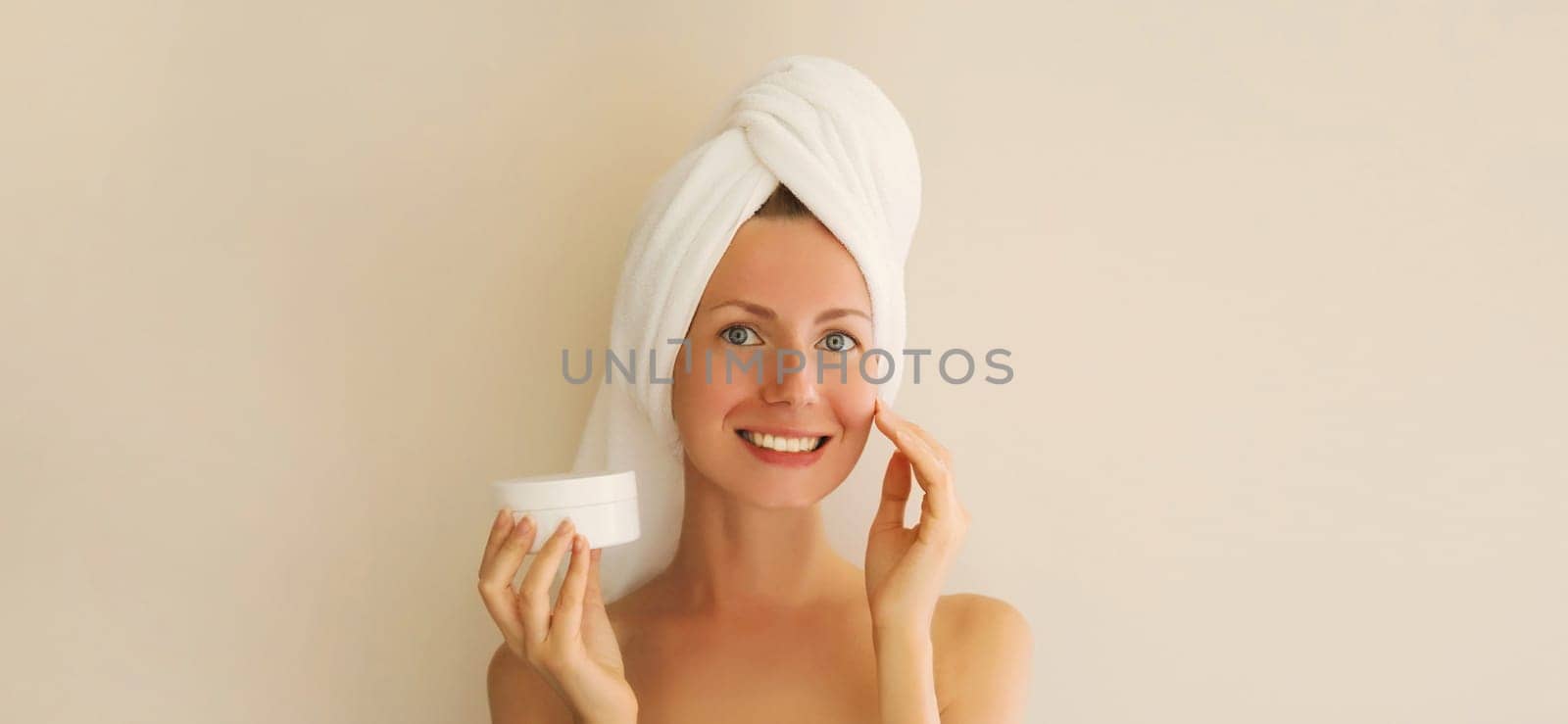 Natural beauty portrait of happy smiling young caucasian woman touches her clean skin applying face cream while drying wet hair with white bath towel on her head