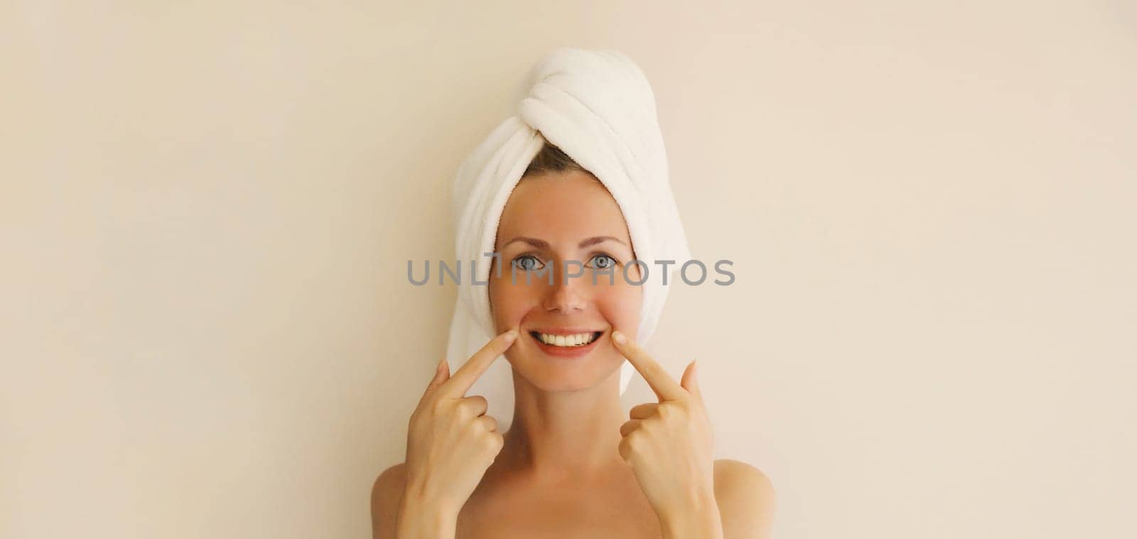 Portrait of happy smiling caucasian young woman points to her white clean teeth while drying her hair with towel on her head after shower in the morning at home