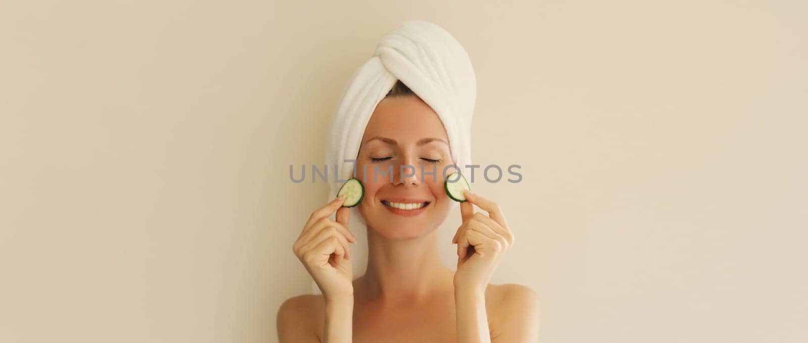 Natural beauty portrait of happy smiling young caucasian woman covers her clean skin holds slices of cucumber drying hair with white bath towel on her head at home