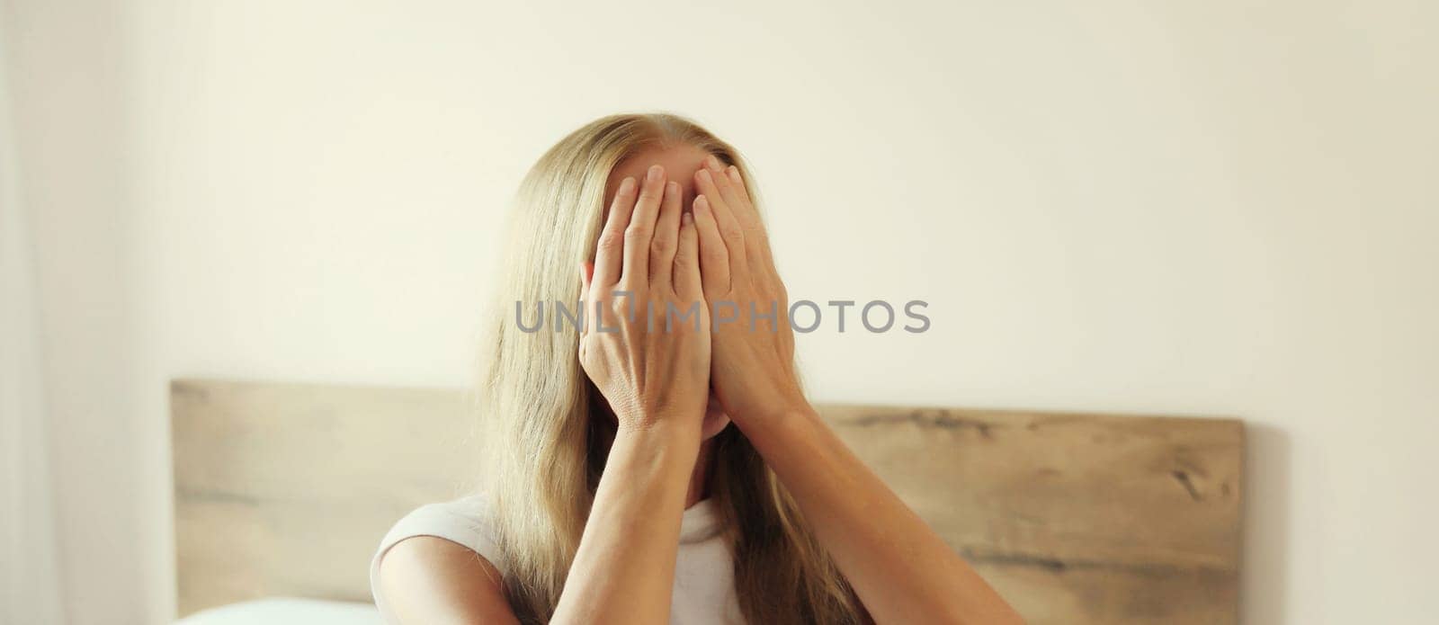 Upset sad crying young woman covering her eyes and face with her hands while experiencing depression, stress, fatigue, mental suffering after sleeping sitting on the bed at home