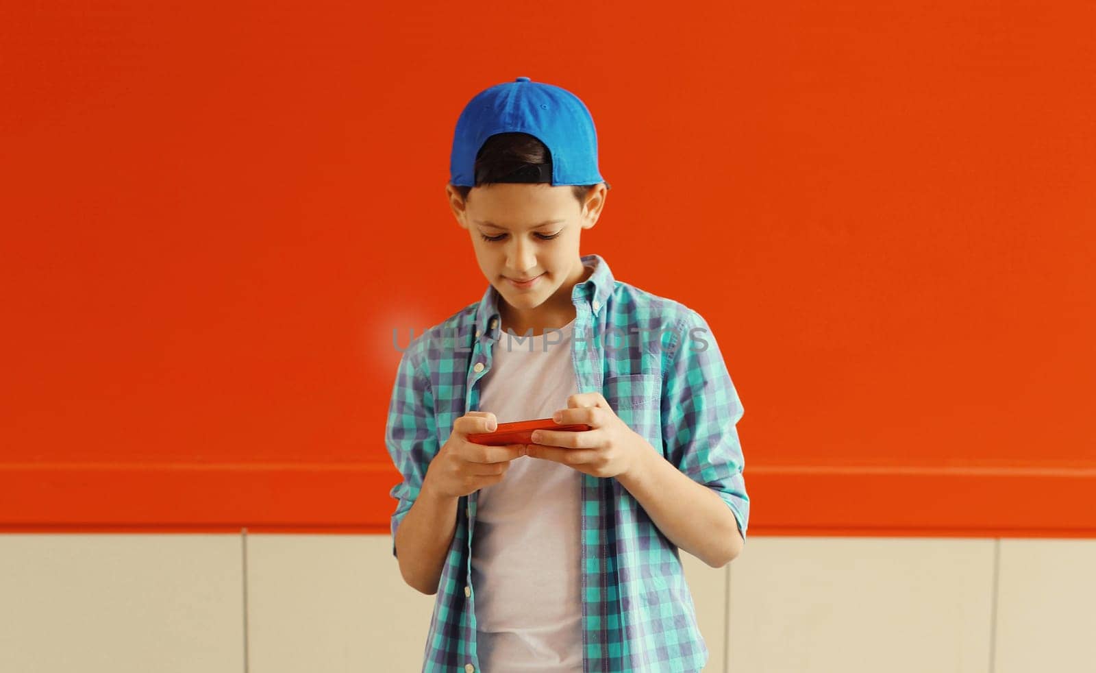 Happy modern teenage boy with phone, kid holding smartphone in the city on colorful red wall background