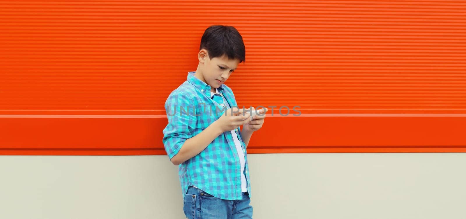 Happy modern teenage boy with phone, kid holding smartphone in the city on colorful red wall background