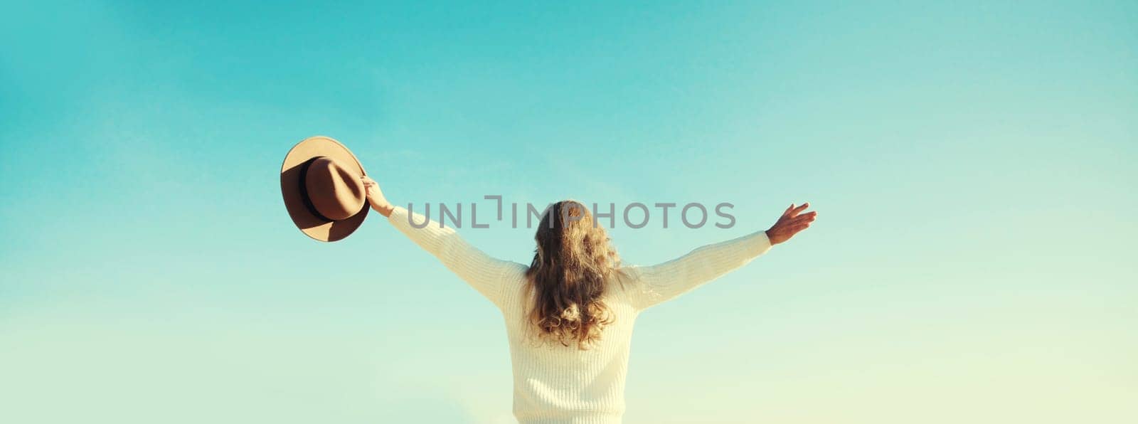 Back view of happy young woman raising her hands up on sunny beach, enjoying amazing landscape, warm weather, wear hat, white sweater, standing on sea coast, nature background