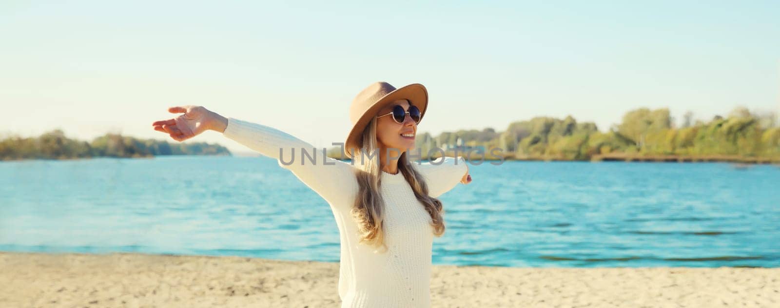 Happy smiling woman traveler raising her hands up on beach, enjoying warm weather, wear hat, white sweater, standing on sea coast, blue sky, nature background