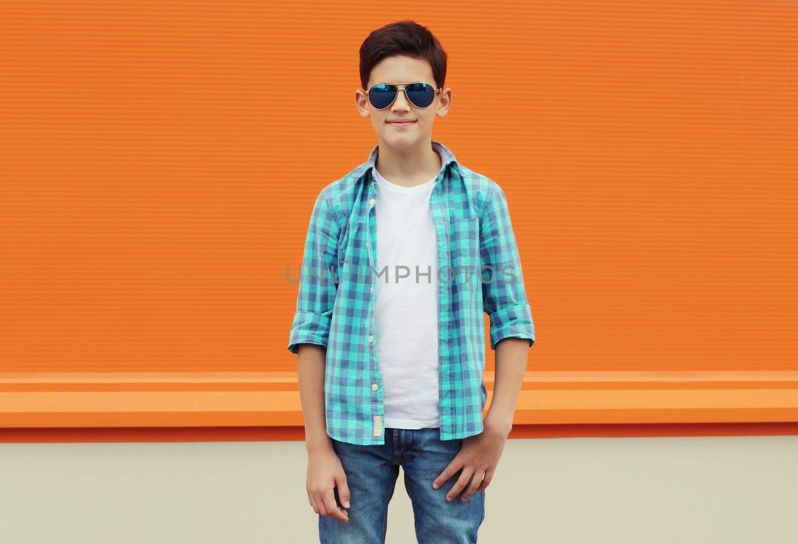 Modern teenage kid boy in casual clothes with glasses standing on city street, orange wall background