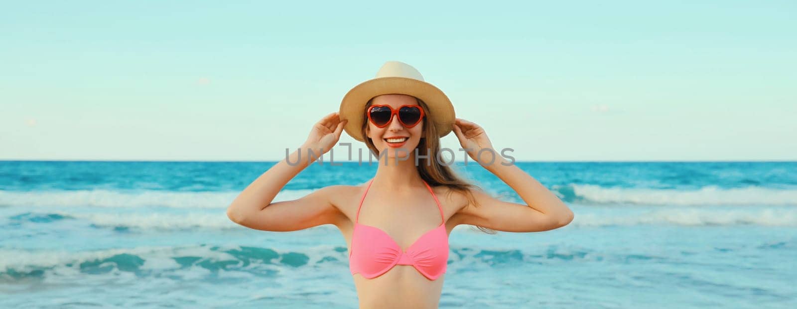 Summer vacation, beautiful happy lovely woman in pink bikini swimsuit, tourist straw hat, red heart shaped glasses on the beach on sea coast with waves