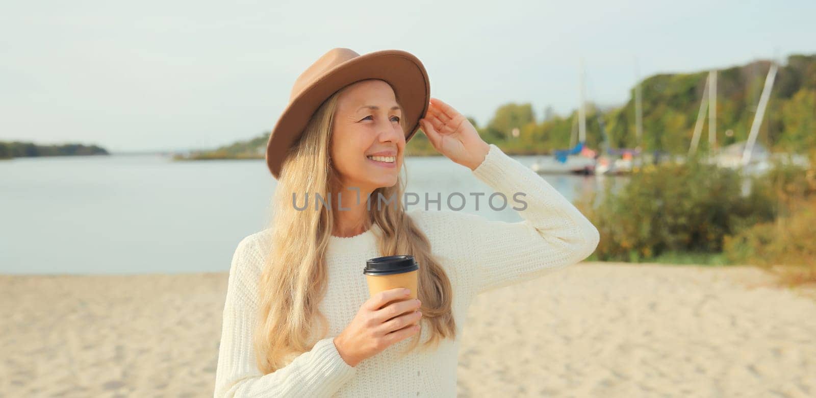 Stylish happy woman with cup of coffee drink standing on beach, smiles, looking away, wear hat, white sweater, on sea coast background