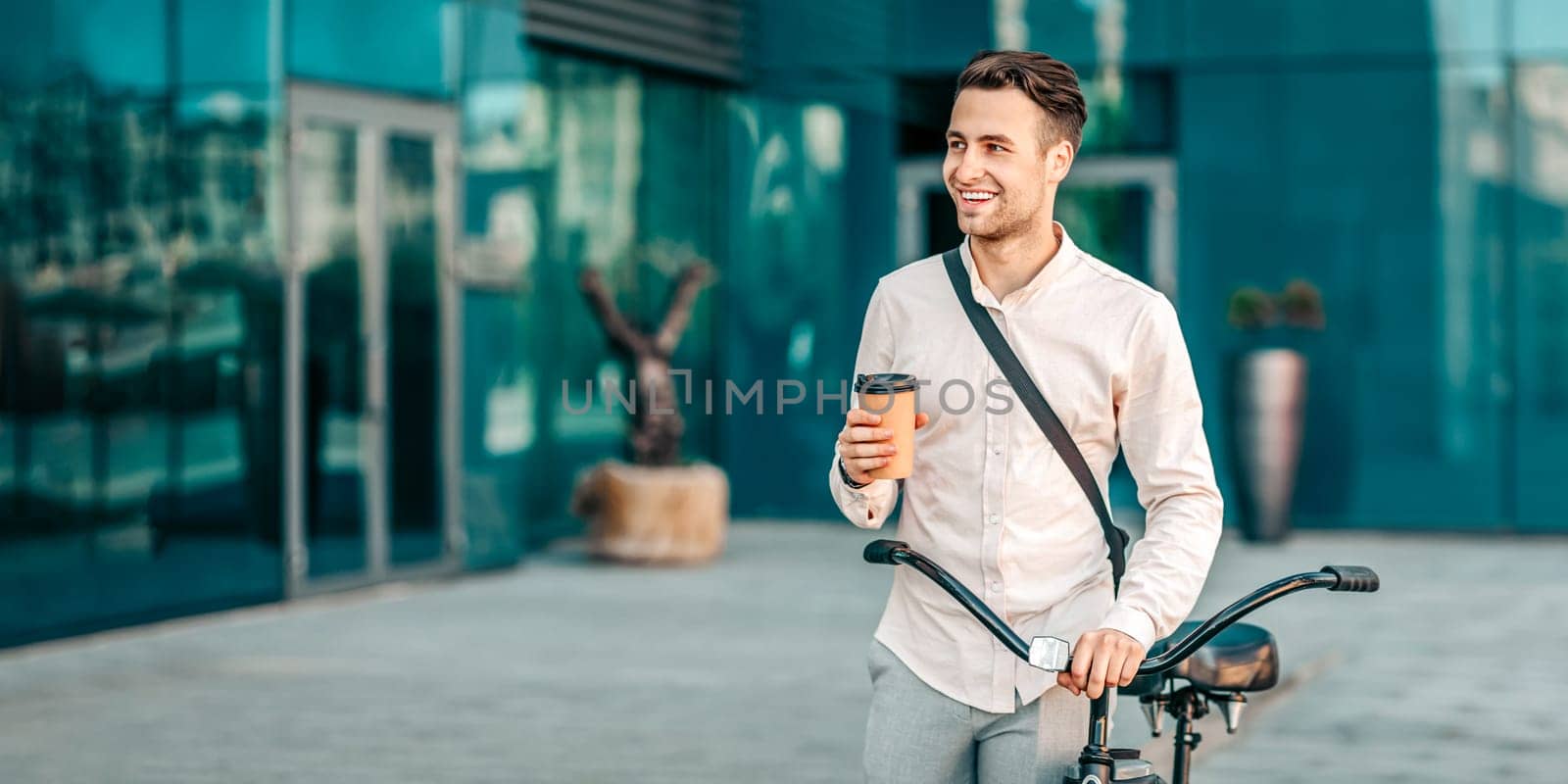 Young man enjoying coffee while standing with bike on urban street, looking at copy space