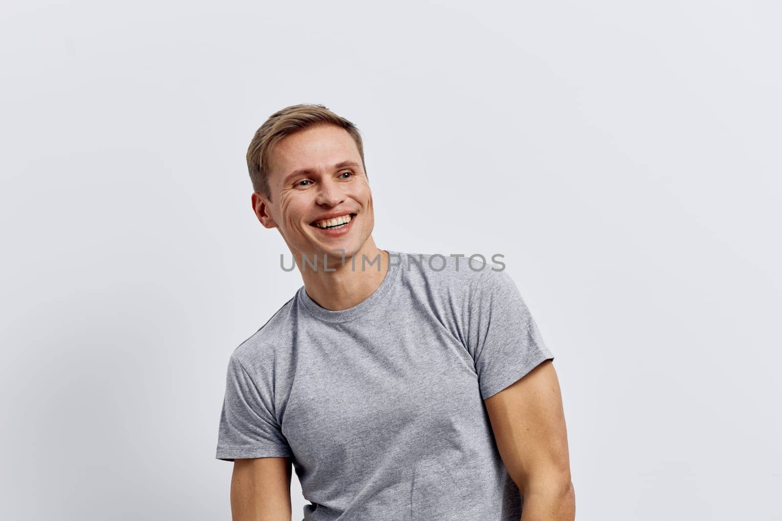 Smiling young caucasian man wearing grey t-shirt looking away seated on plain light background with relaxed posture. Casual lifestyle concept emphasizing digital nomad remote work and freelance flexibility. Concept: digital nomad work