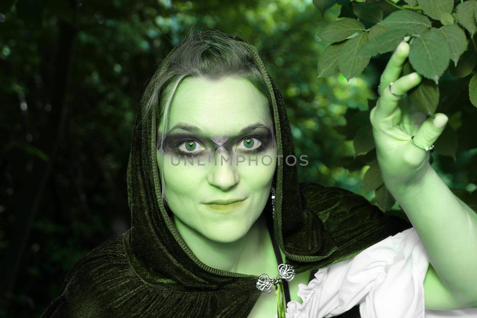 A woman dressed as a green forest spirit or witch looks out of the bushes