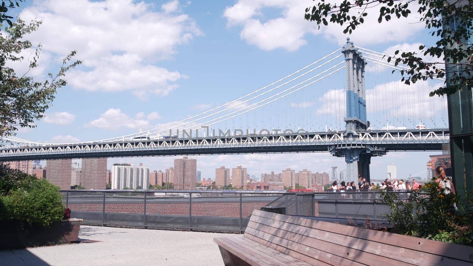 New York City, United States - 30 Aug 2023: Cable-stayed Manhattan Bridge to Brooklyn Dumbo. Bench on Time Out Market rooftop, iconic United States travel destination. People on NYC roof vista point.