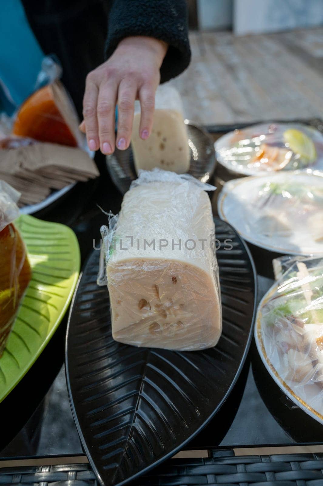 Hand of a seller and a piece of cheese in a transparent package at a farmers' market. Natural cheese at a city fair. A large piece of cheese on a black oval plate.