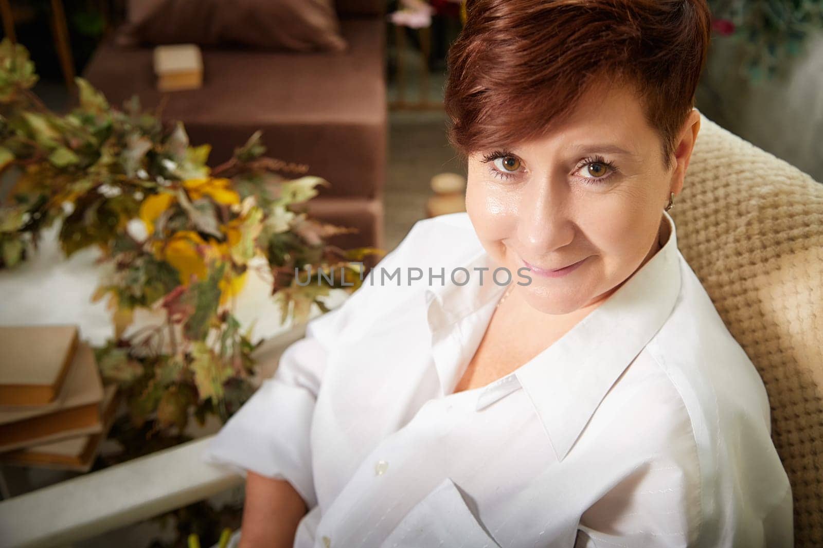 Blonde woman smiles warmly while relaxing in a cozy room filled with plants and soft furnishings, engaged in blogging.