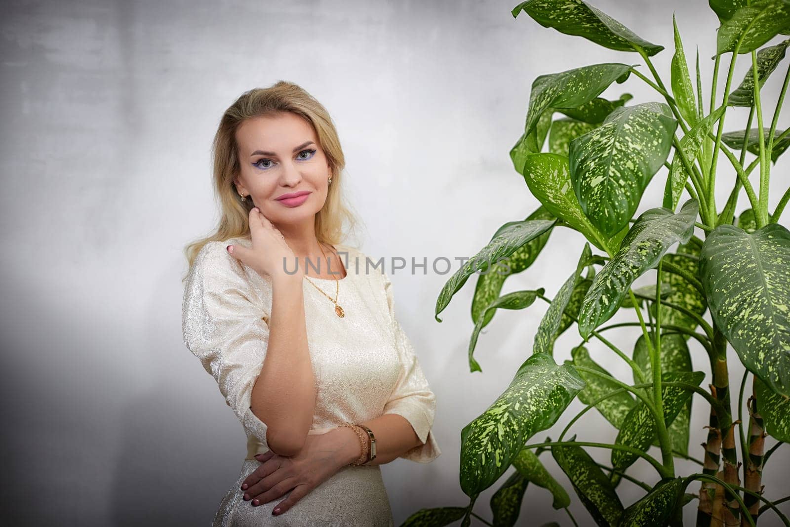 A middle-aged woman styled as a blogger poses elegantly indoors next to a lush plant, showcasing her lifestyle.