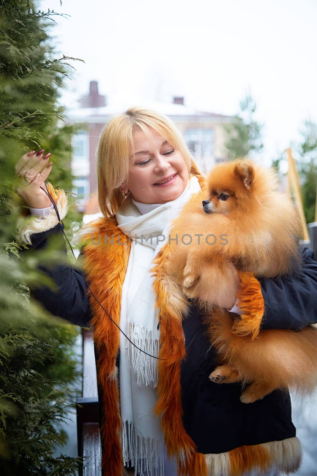 A middle-aged blonde woman lovingly holds her fluffy Pomeranian dog while enjoying a snowy day outdoors.