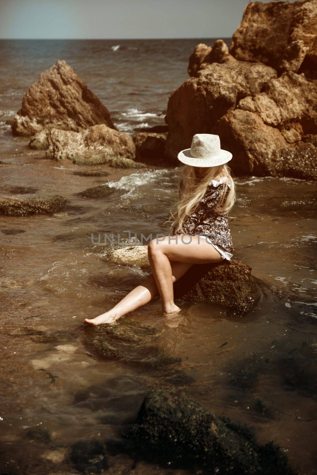 young blonde in short dress and hat sitting on rock in sea on beach. her hair fluttering in wind