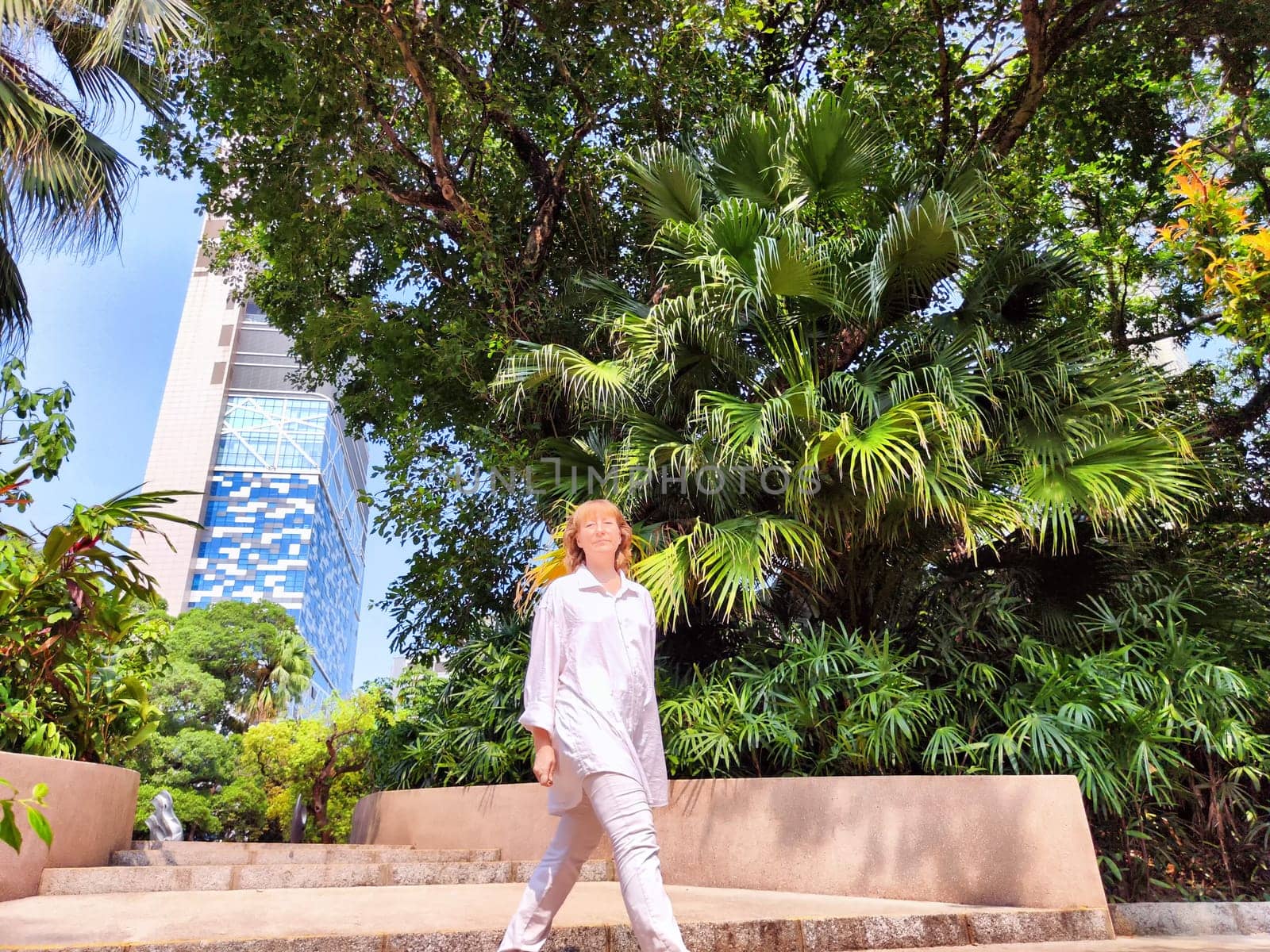 Blonde adult walks confidently through a vibrant garden surrounded by tropical plants and modern buildings on a sunny day.