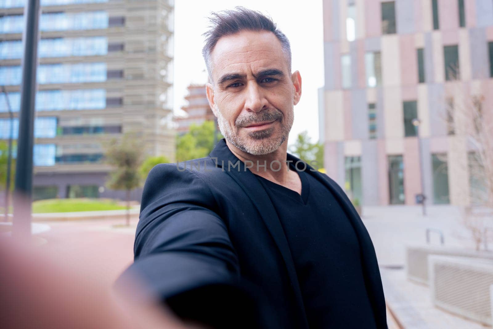 Caucasian man with confident expression takes a selfie in a modern city environment. Wearing black outfit, he stands among tall buildings. Urban lifestyle and personal branding concept.