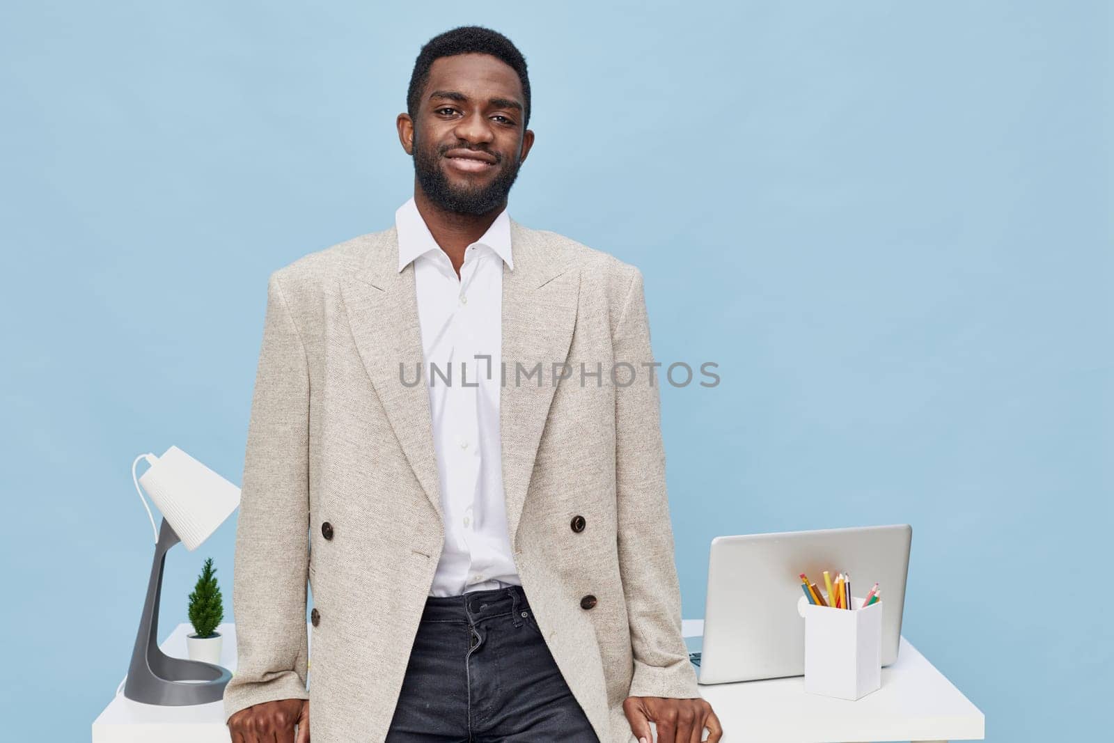 A cheerful man in a beige blazer and white shirt standing at his desk with a laptop, desk lamp, and writing utensils, captured in a bright modern office with a light blue background, conveying professionalism and approachability. Concept: modern workplace wellness