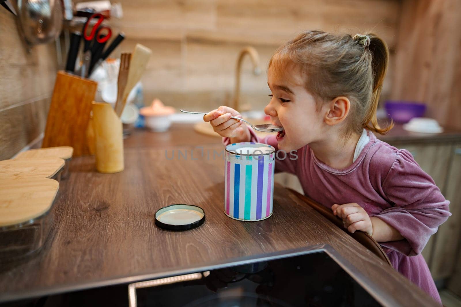 A cheerful little girl sits at a wooden kitchen counter, eagerly tasting her dessert from a colorful container. The warm ambiance radiates happiness, enhancing her playful spirit.