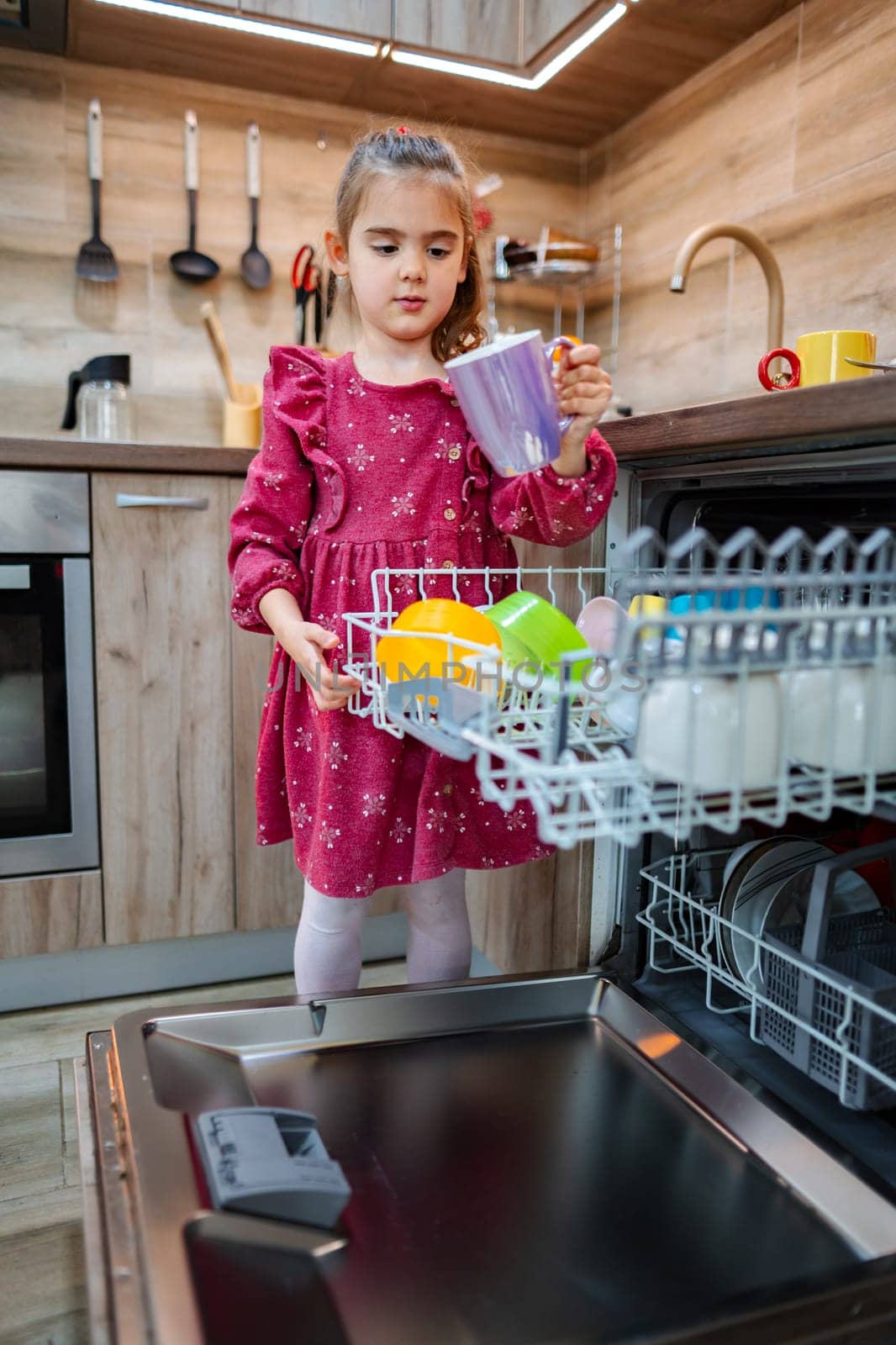 A young child in a bright pink dress carefully loads colorful dishes into a dishwasher in a modern kitchen. Soft light illuminates the wooden cabinetry, highlighting her focused expression.