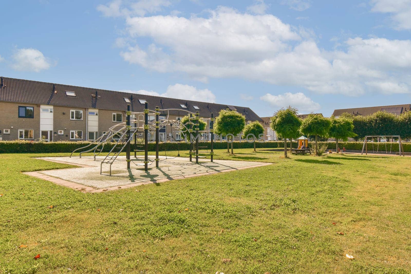 A vibrant playground featuring climbing structures surrounded by well-maintained grass, trees, and residential buildings under a clear blue sky.
