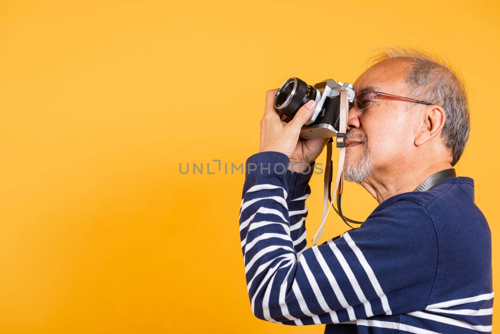 Portrait Asian old man wearing glasses look viewfinder vintage SLR camera studio shot isolated yellow background, smiling happy Photographer elderly man gray haired taking a picture of himself