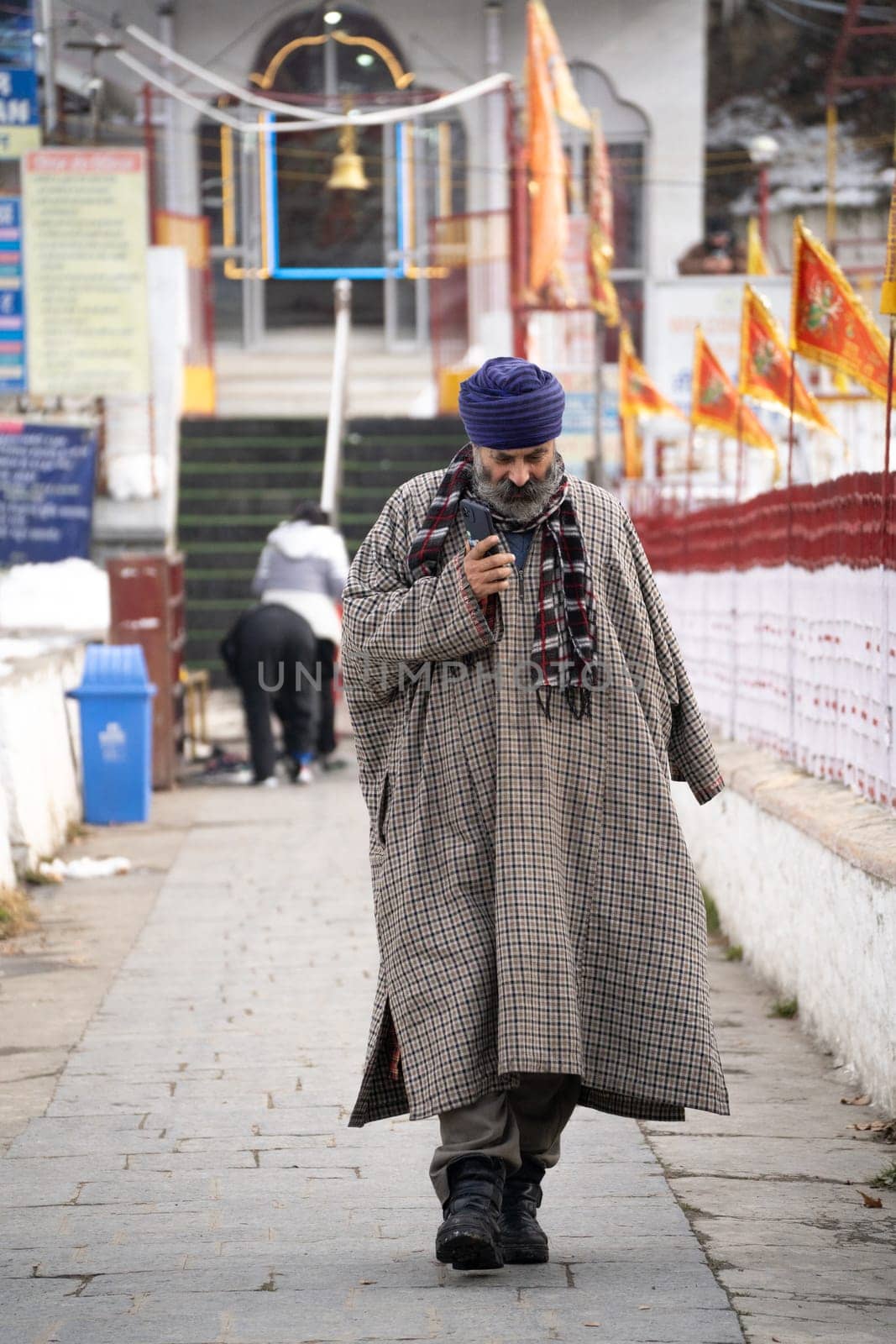 Srinagar, Kashmir - 4th Jan 2025: Sikh man wearning the traditional phiran overcoat cloak of kashmir walking out of a temple while talking on phone