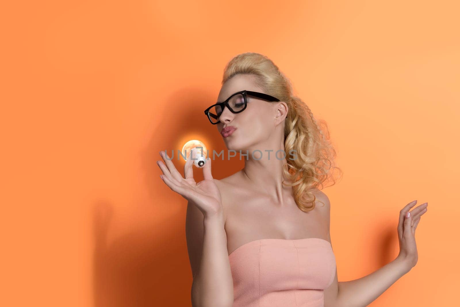 funny portrait of blonde girl with pink dress and black glass. She kissing  light bulb on orange background