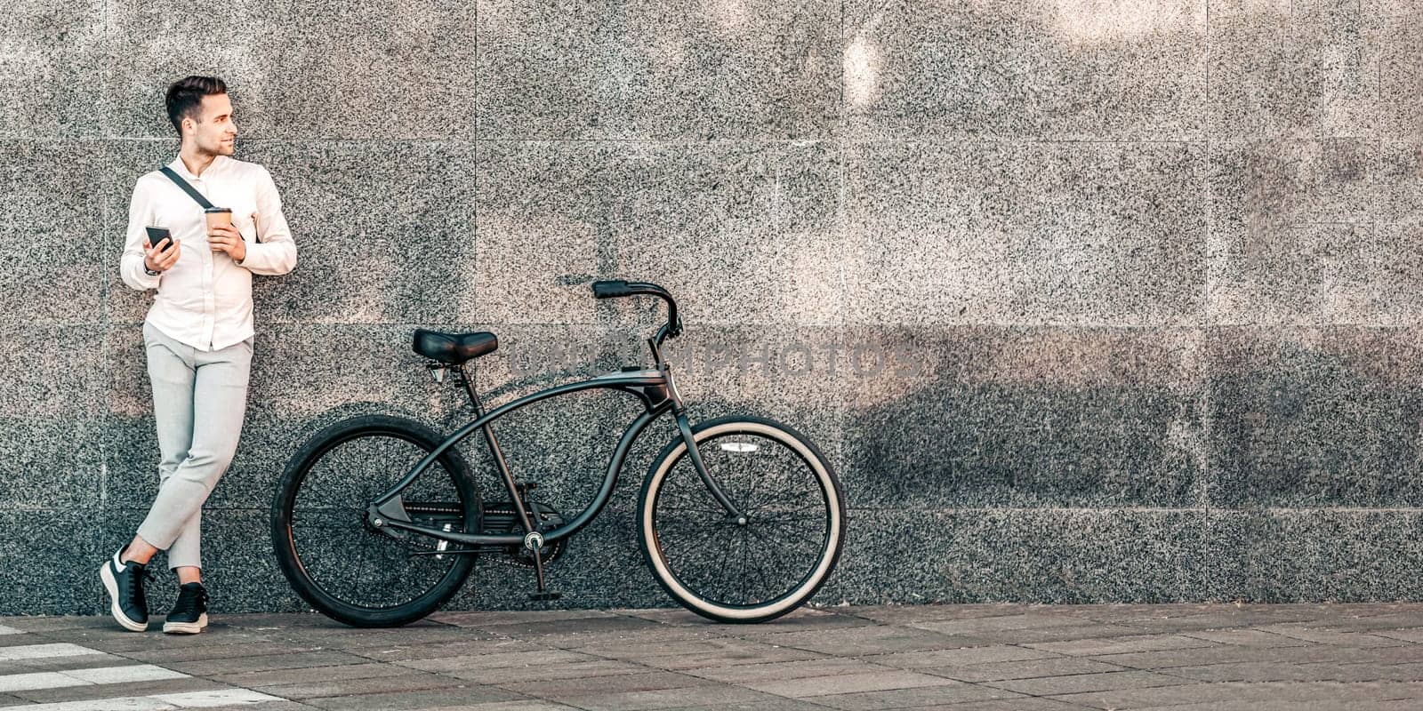 A young man leans against textured concrete wall, casually holding his smartphone while his stylish bike rests nearby. This vibrant street captures a millennial lifestyle in urban scenery, copy space
