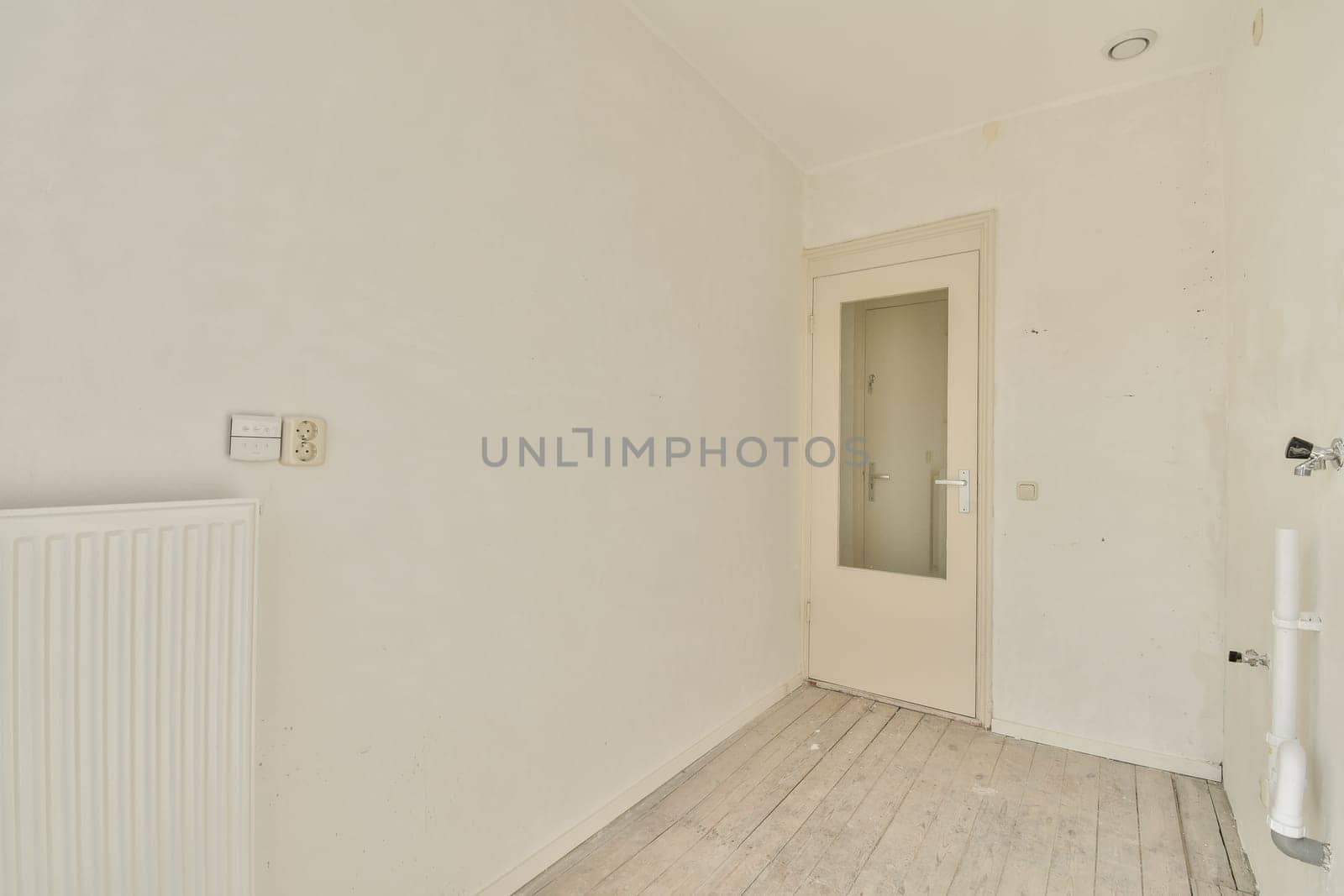 A clean and empty interior space featuring white walls, a light wooden floor, and a simple door. The ambiance conveys a sense of openness and simplicity.