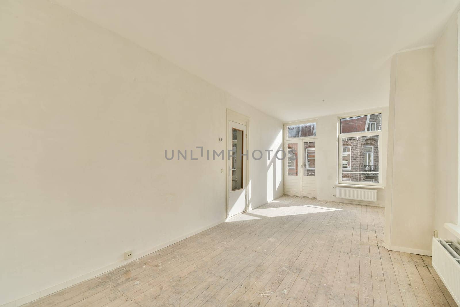 A bright, spacious room featuring large windows that allow abundant natural light. The area has a minimalistic design with light-colored walls and wooden flooring.