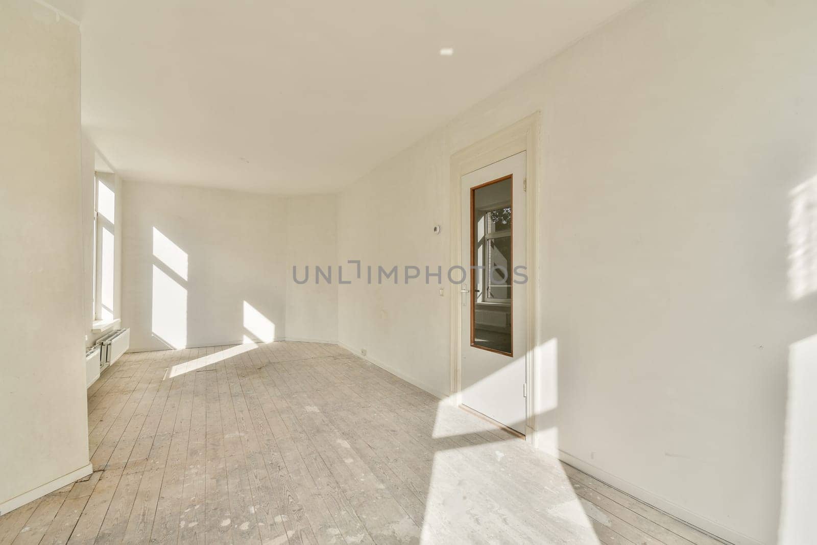 A spacious, empty room featuring pristine white walls and wooden flooring, illuminated by natural light streaming in through large windows.