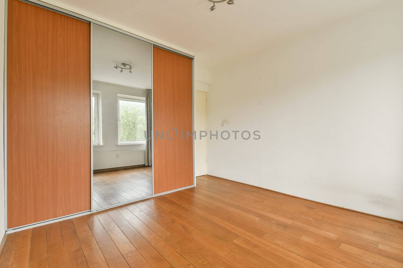 A spacious minimalist room featuring a large mirrored wardrobe and warm wooden flooring. The natural light filters through the window, creating a serene atmosphere.
