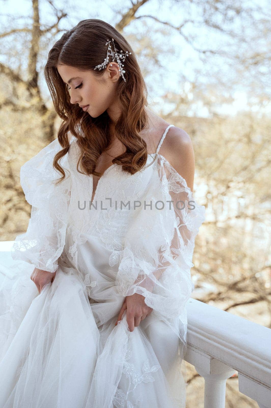 Young brunette bride in traditional long white dress posing on hotel balcony in spring. white dress with long sleeves, corset and open shoulders. Bride portrait, wedding makeup, fashionable bride