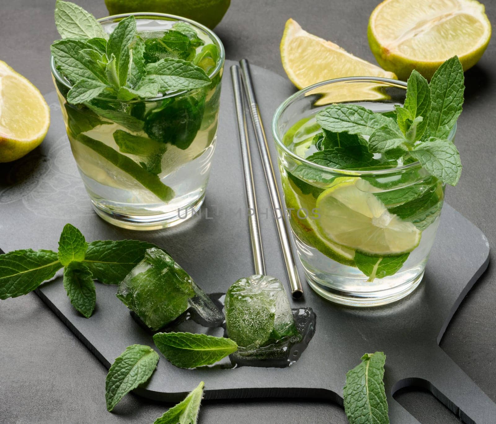 Glass of mojito cocktail with fresh mint leaves, lime slices, and ice cubes on a dark stone background. Refreshing summer drink with stainless steel straws