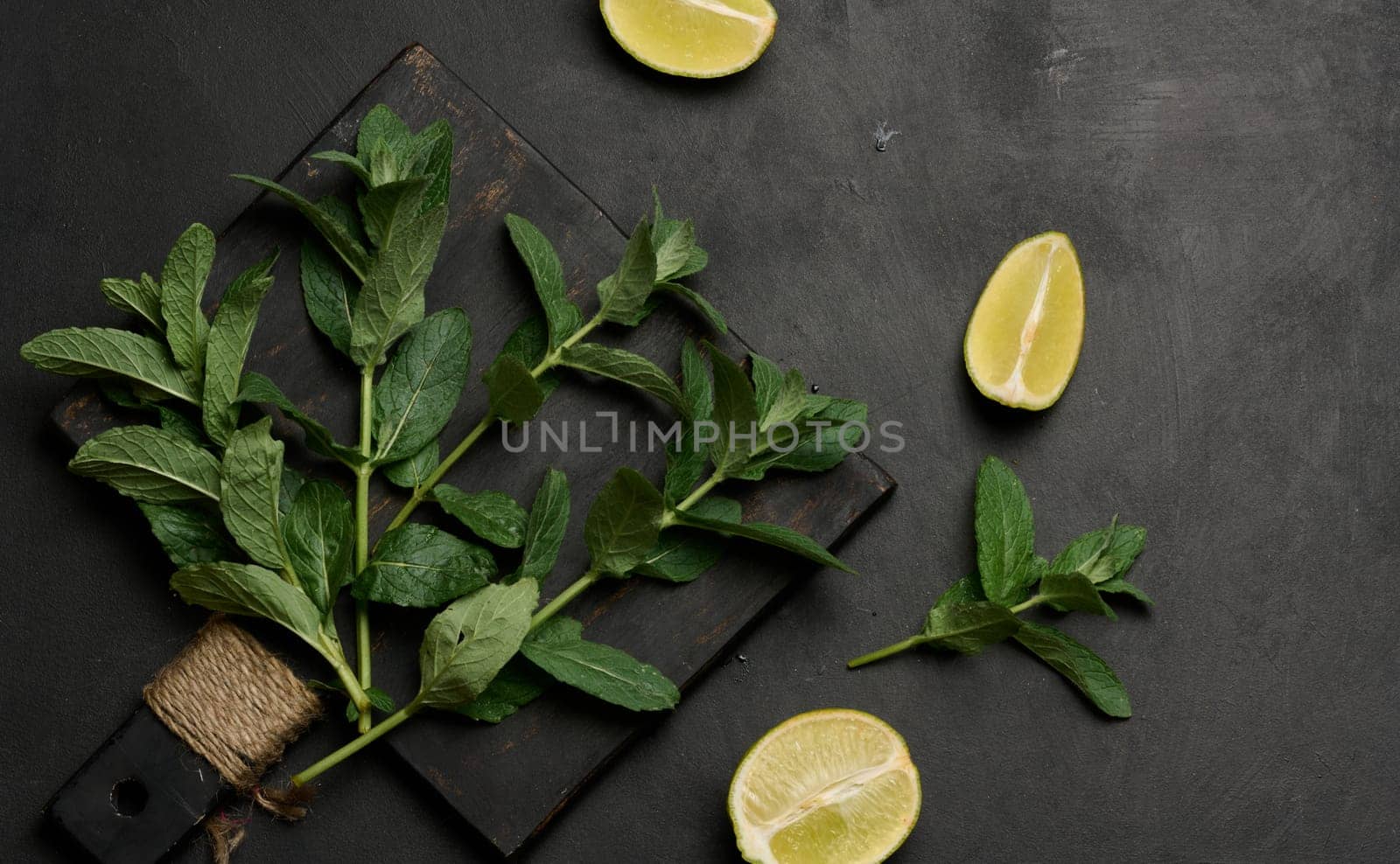 Whole and cut limes with fresh mint leaves arranged on a dark textured surface. Composition perfect for food, drink, or summer themes